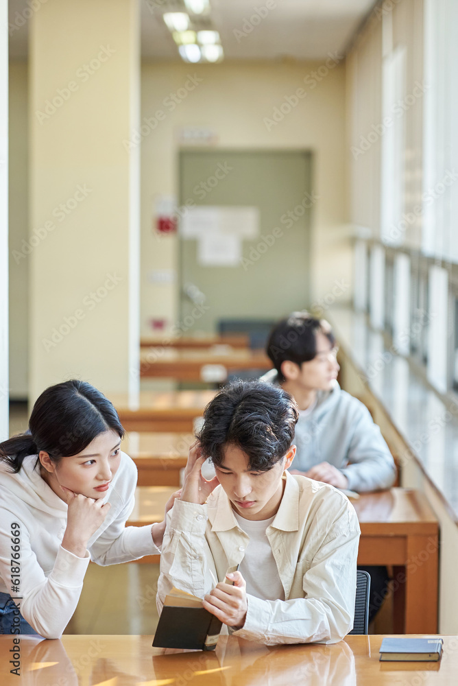 Three college students, both male and female, are sitting or standing at a desk in a library at a South Korean university in Asia, looking at laptops and books while having a conversation. Three college students, both male and female, are sitting or standing at a desk in a library at a South Korean university in Asia, looking at laptops and books while having a conversation.