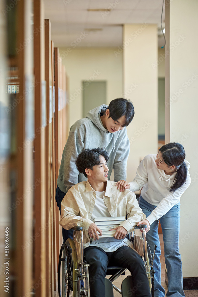 A young male model with a disability sitting in a wheelchair reading a book in a university library in Asia, with two young male and female college students helping him A young male model with a disability sitting in a wheelchair reading a book in a university library in Asia, with two young male and female college students helping him
