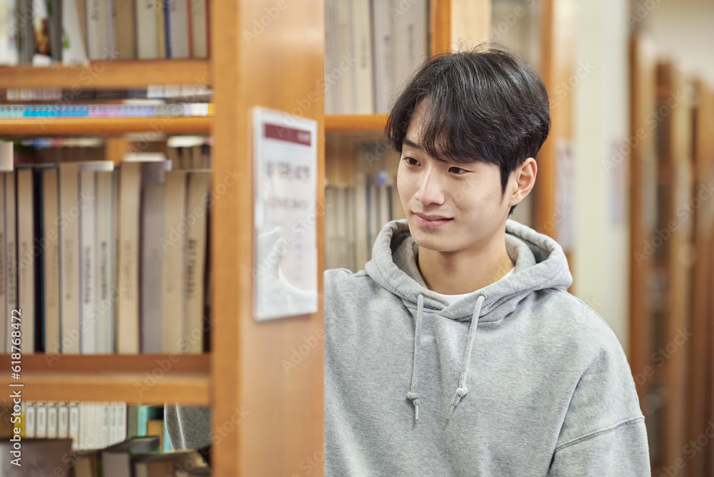 A young man attending college is browsing and reading books on the shelves with different expressions in his university library in South Korea, Asia. A young man attending college is browsing and reading books on the shelves with different expressions in his university library in South Korea, Asia.
