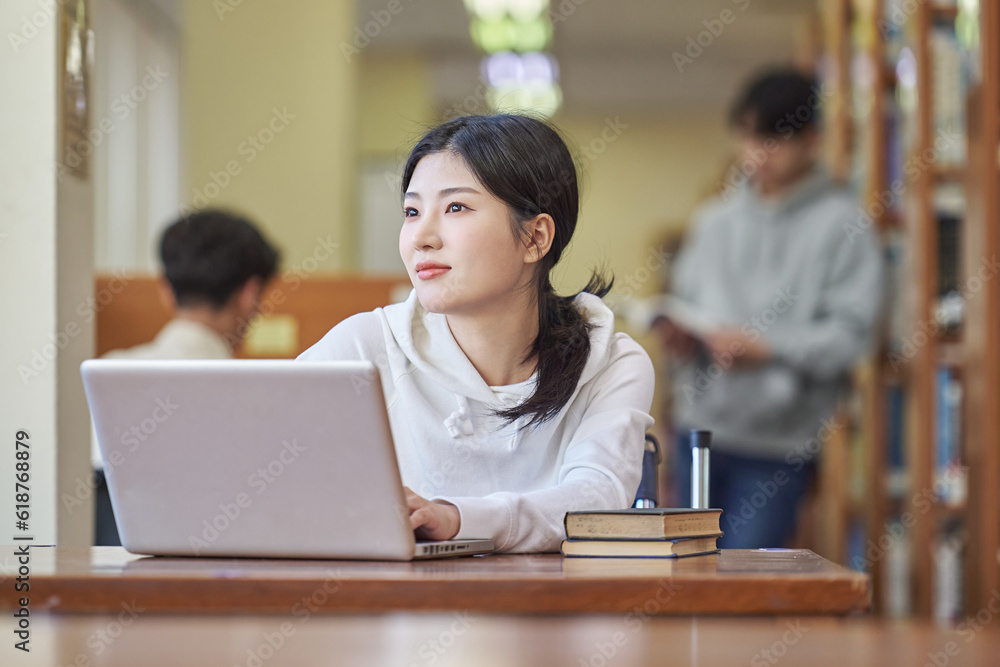 Young Asian Korean female model in library looking at laptop or book, lecture, assignment, discussion, male model in background, bookshelf in background Young Asian Korean female model in library looking at laptop or book, lecture, assignment, discussion, male model in background, bookshelf in background