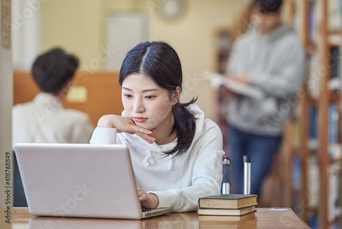 Young Asian Korean female model in library looking at laptop or book, lecture, assignment, discussion, male model in background, bookshelf in background