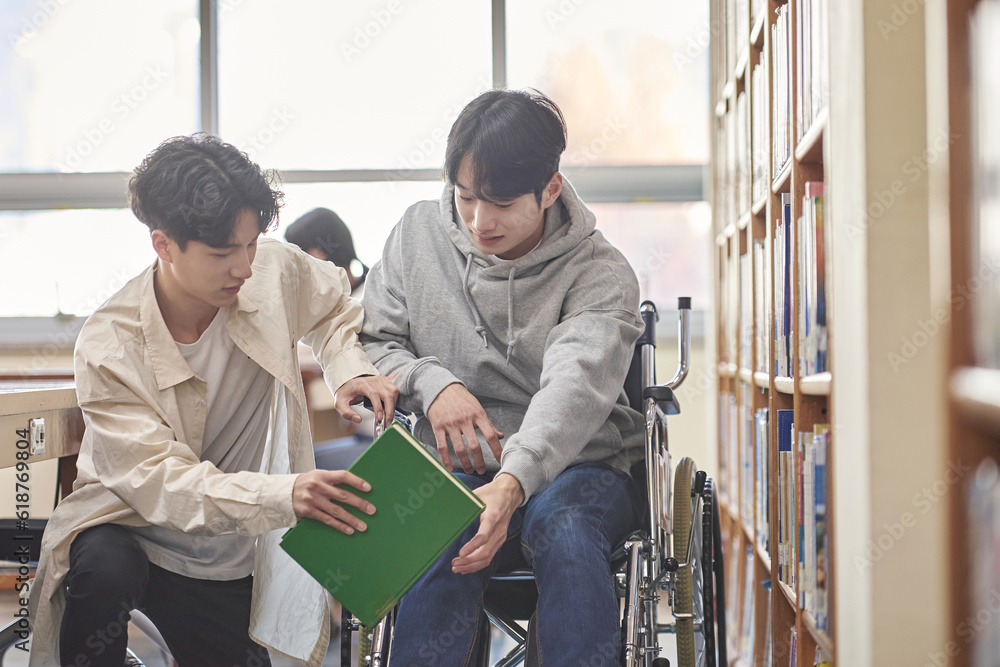 Young handicapped disabled male college student model in wheelchair looking for books in library bookshelf at university in Asia, helping male college student Young handicapped disabled male college student model in wheelchair looking for books in library bookshelf at university in Asia, helping male college student