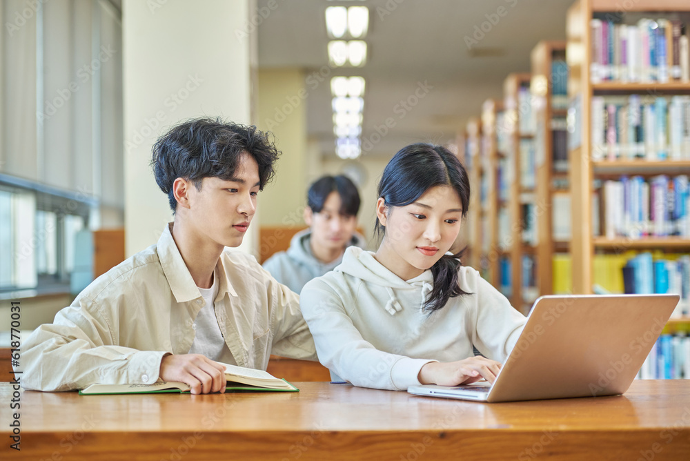 Two young college student man and woman couple model and solo male model looking at laptop and book together in library of Asian Korean university Two young college student man and woman couple model and solo male model looking at laptop and book together in library of Asian Korean university