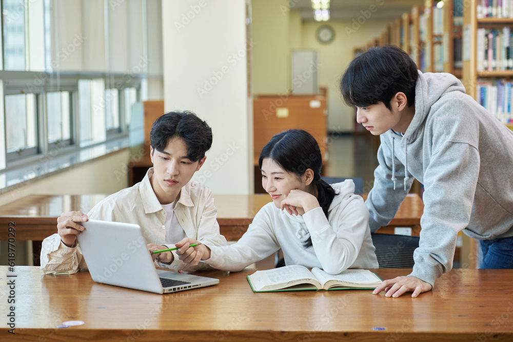 Two young college student man and woman couple model and solo male model looking at laptop and book together in library of Asian Korean university Two young college student man and woman couple model and solo male model looking at laptop and book together in library of Asian Korean university