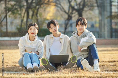 Three young college student male and female models sitting on the lawn at the university in autumn in Asian Korea, watching a laptop, listening to a lecture, discussing or talking