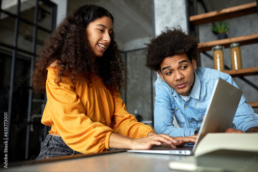 Side view of charming young black happy smiling female with curly hair sitting at laptop playing video game or gambling online while her afro boyfriend looking at screen with shocked puzzled face Side view of charming young black happy smiling female with curly hair sitting at laptop playing video game or gambling online while her afro boyfriend looking at screen with shocked puzzled face