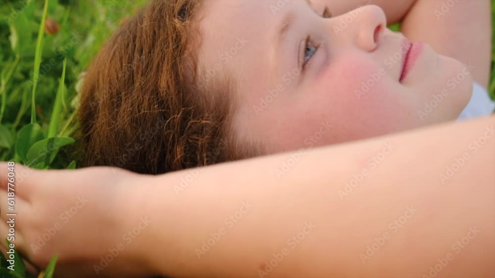 Close up side view of happy cute little girl lying on green grass in summer warm day. Portrait adorable child is dreaming looking at sky on lawn outdoors. Children day. Childhood and nature concept Close up side view of happy cute little girl lying on green grass in summer warm day. Portrait adorable child is dreaming looking at sky on lawn outdoors. Children day. Childhood and nature concept