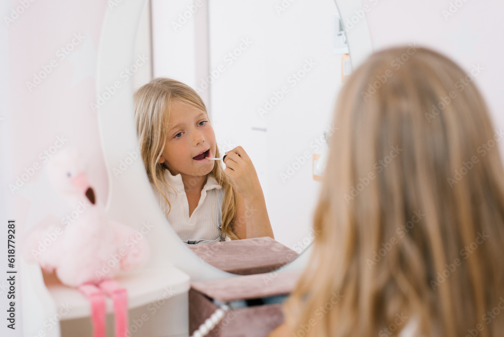 Child girl paints her lips with glitter or balm in front of a mirror in her room Child girl paints her lips with glitter or balm in front of a mirror in her room
