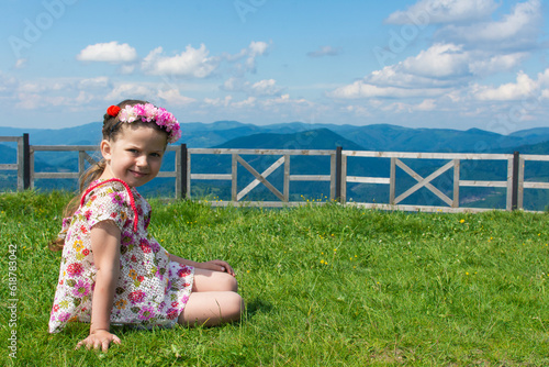 A smiling caucasian girl sits with a wreath on her head on a grass high in the mountains on a summer sunny day in Ukraine.