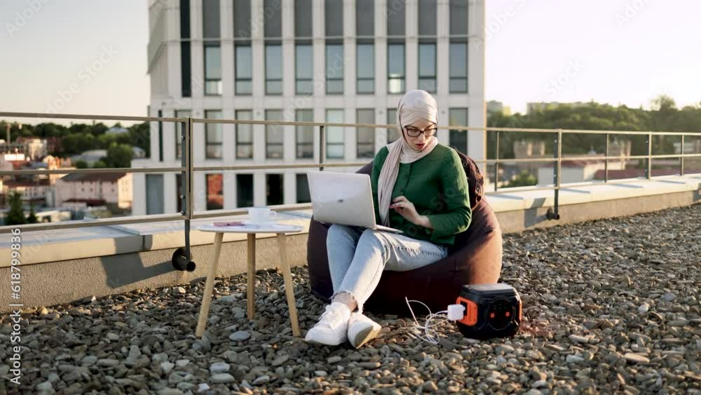 Wide view of arabian woman in traditional headscarf working on laptop while sitting in soft chair in fresh air. Discharged gadget being connected to renewable energy source on panoramic terrace. Wide view of arabian woman in traditional headscarf working on laptop while sitting in soft chair in fresh air. Discharged gadget being connected to renewable energy source on panoramic terrace.