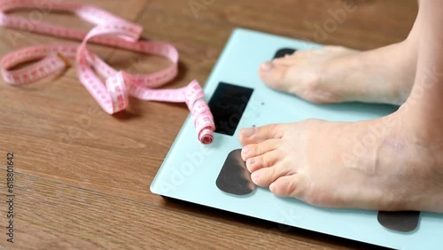 White floor scales and women's feet standing on them. A tailor's centimeter or measuring tape lying nearby to measure body changes. The concept of a healthy lifestyle and body weight tracking