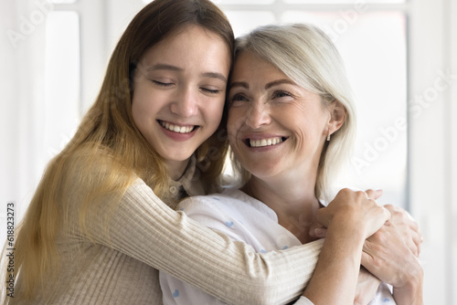 Cheerful teen granddaughter kid and happy grandmother enjoying warm family moment, meeting at home, hugging with toothy smiles and face touch, celebrating mothers day