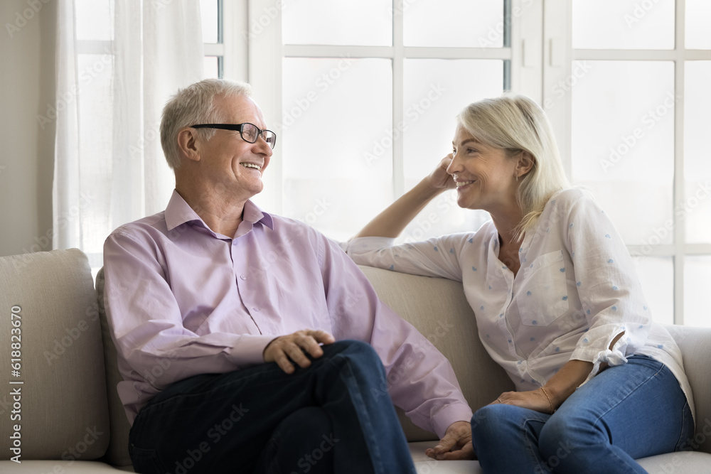 Happy senior dating couple resting on couch at home, talking, laughing, discussing retirement. Mature pretty blonde wife and old husband, speaking, smiling, enjoying relationship