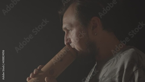 A hipster man plays the Australian aboriginal wind musical instrument Didgeridoo on a dark background. Music footage photo