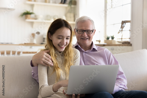 Happy teenager kid and elder grandfather enjoying modern internet technology at home together, girl using application on laptop computer, teaching granddad to use online service