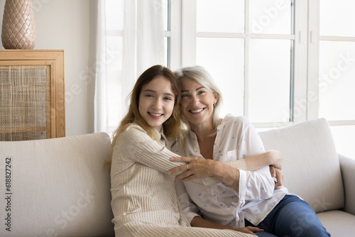 Beautiful grandma and teenager granddaughter posing at home, sitting on couch together, hugging, enjoying shooting, family leisure, hugging, looking at camera, smiling, laughing © fizkes