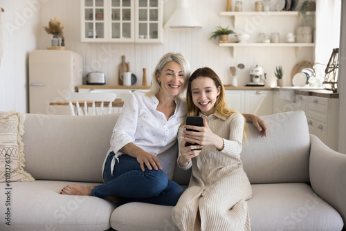 Cheerful mature granny and young teenage kid girl using media application on smartphone together, enjoying Internet technology, family communication, talking selfie photo