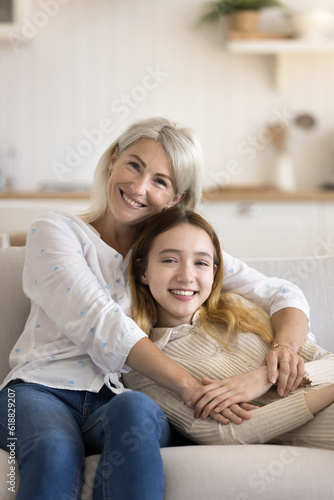 Happy mature grandma and adorable teenage grandkid girl resting on home couch, hugging with love, care, tenderness, looking at camera, smiling, relaxing on family meeting. Vertical shot portrait