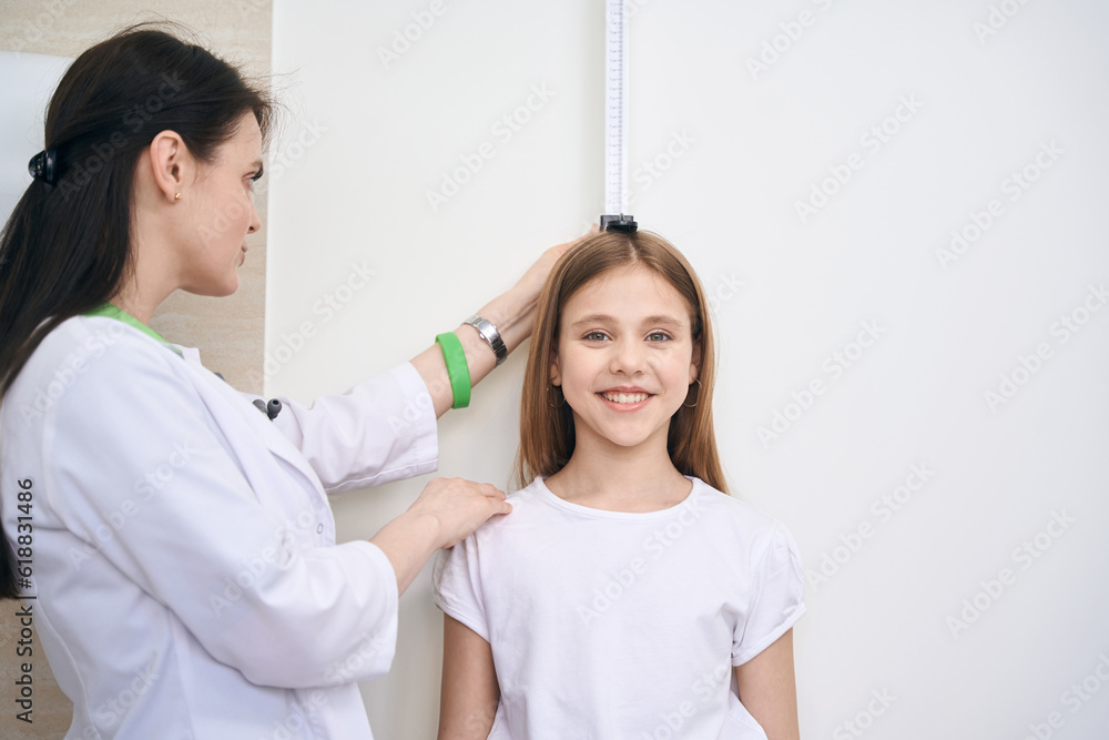 Woman nurse checking little girl height using special measuring plank - obrazy, fototapety, plakaty 