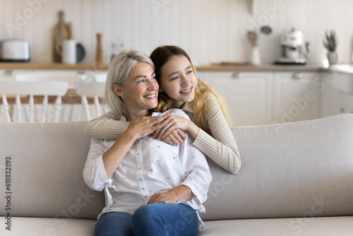 Positive dreamy teenage kid hugging mature grandma from behind at home, looking away, smiling, thinking, dreaming, discussing future plans, enjoying family relationships, bonding, affection
