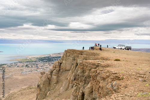 Un grupo mediano de personas se encuentran en la cima del Cerro Huyliche mirando el paisaje de la ciudad de El Calafate Argentina junto a dos vehículos todo terreno. photo