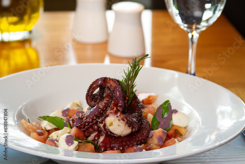 Octopus tentacle served with tomato, cheese, basil and sauce on a white plate on a table with a glass of wine, oil, salt and pepper. Appetizing salad plate. Meal concept