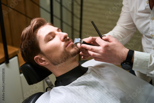 Man cuts a beard to a young client in a barbershop