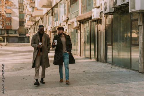 Young white businessman walking and talking business to his black male partner. They are holding cups of hot morning coffee