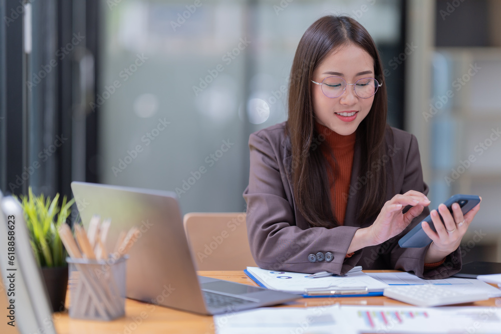 Sharing good business news. Attractive young businesswoman talking on the mobile phone and smiling while sitting at her working place in office and looking at laptop PC.