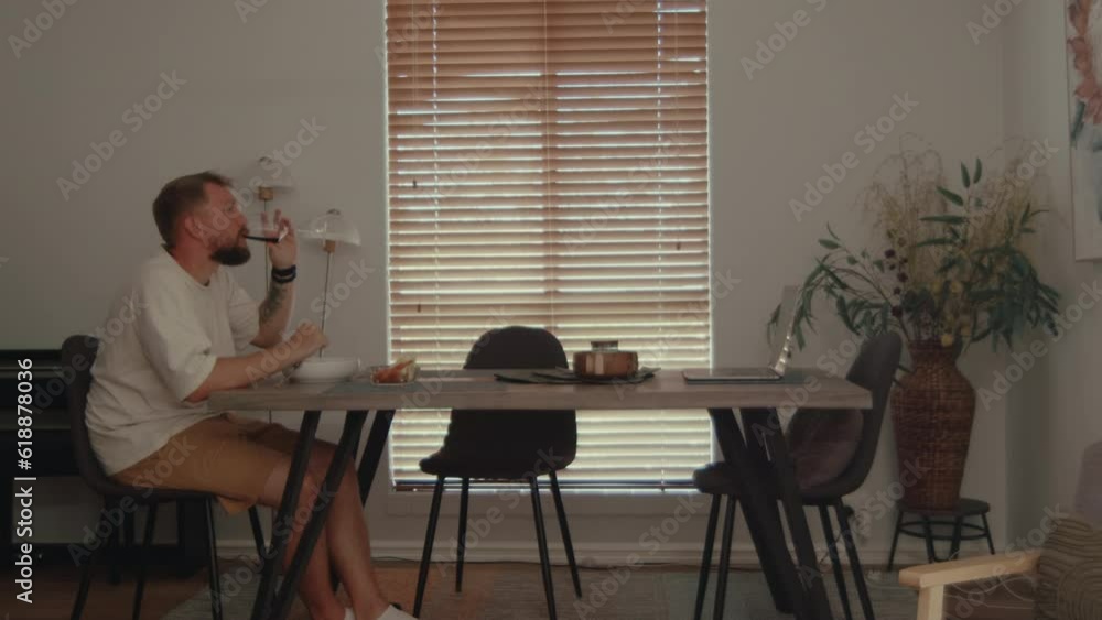 Young man sits at a set table, enjoying a delicious dinner with a glass of wine in a cozy dining room. He is engrossed in captivating content on his laptop, watching tv-show or online content. Young man sits at a set table, enjoying a delicious dinner with a glass of wine in a cozy dining room. He is engrossed in captivating content on his laptop, watching tv-show or online content.