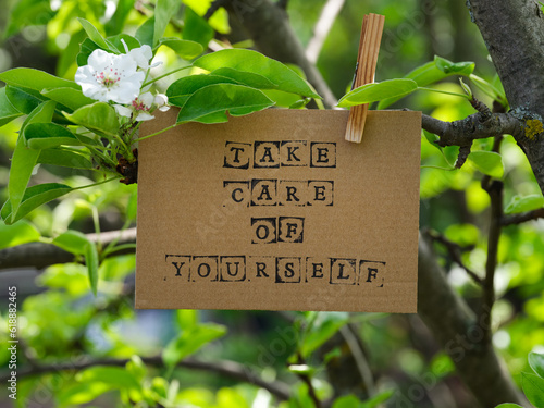 Piece of cardboard with words Take Care of Yourself on it hanging on a pear tree branch with blossoms and leaves using a wooden clothespin. © rosinka79