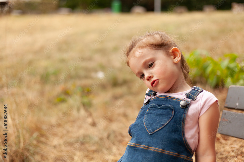 A beautiful blonde little girl with a tail sits on a bench in the park in a denim overalls A beautiful blonde little girl with a tail sits on a bench in the park in a denim overalls