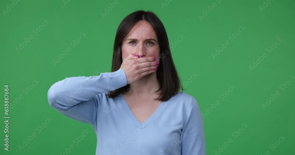 Caucasian woman stands on a green chroma key background, making a keep quiet hand gesture with a serious expression on her face. She covers her mouth with her hand. Caucasian woman stands on a green chroma key background, making a keep quiet hand gesture with a serious expression on her face. She covers her mouth with her hand.