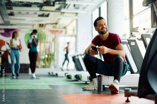 Young athletic man using mobile phone while relaxing after sports training in gym.