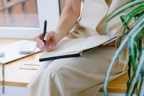 Woman in a beige trouser suit sits on the windowsill in her office and takes notes in a notebook. Business woman plans her schedule in a diary