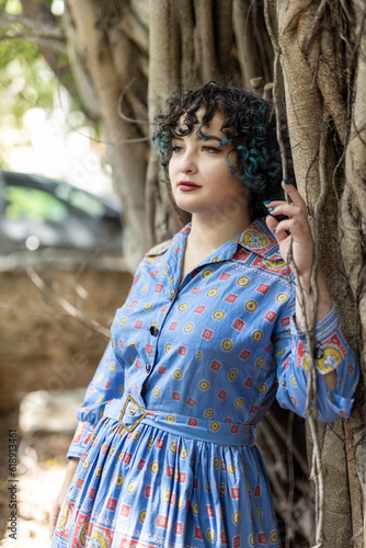 Smiling brunette girl twenty-five years old dressed in a vintage blue patterned dress with a small purse bag leaned against the trunk of a large ficus in the park