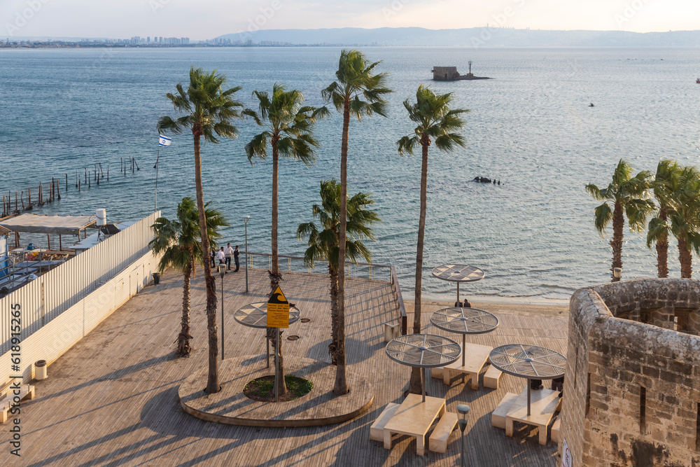 Naklejka premium Acre, Israel - January 08, 2023: Sunset view of the Horses beach, the sea wall and the fishing port, with locals and visitors, in the old city of Acre (Akko), Israel
