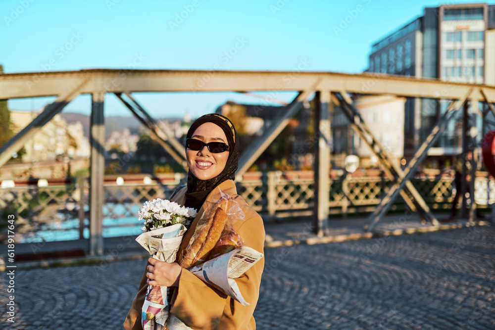 Fototapeta premium A hijab woman in stylish sunglasses and an elegant French outfit, walking through the city at sunset, carrying a bouquet, bread, and newspaper, radiating a sense of cultural charm and serenity