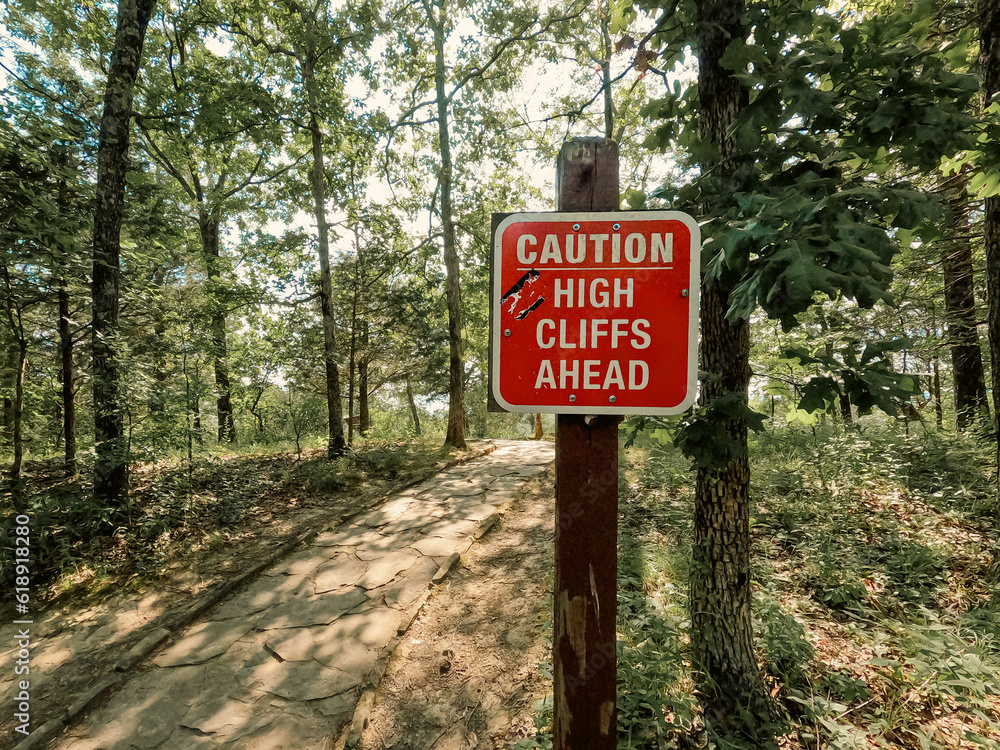 Caution High Cliffs Ahead Trail Sign. Red sign post cautioning visitors that high cliffs are ahead. Forested woodlands with tall green leafy trees are in the background of view. Caution High Cliffs Ahead Trail Sign. Red sign post cautioning visitors that high cliffs are ahead. Forested woodlands with tall green leafy trees are in the background of view.