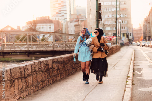 Wallpaper Mural Couple woman one wearing a hijab and a modern yet traditional dress, and the other in a blue dress and scarf, walking together through the city at sunset. One carries a bouquet and bread, while the Torontodigital.ca