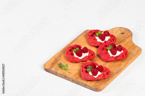 Red heart-shaped waffles with cream cheese, raspberries and micro greenery. Waffles with the addition of beet juice. On a serving wooden board. White background. Valentine's Day. Copy space
