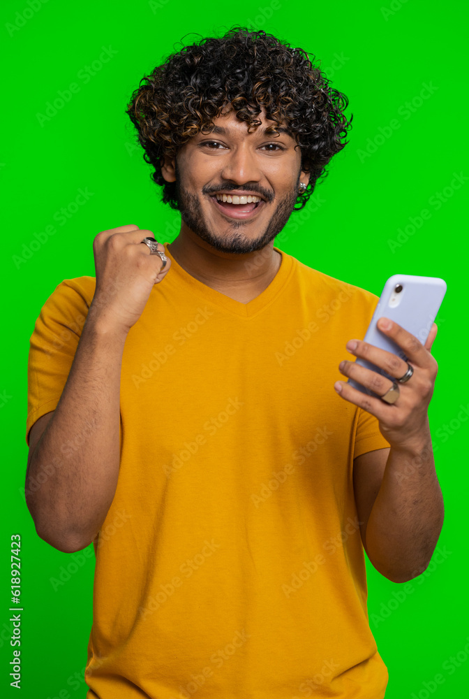 Happy excited joyful indian man in green shirt use smartphone typing browsing shouting say wow yes found out great big win, good news, lottery goal achievemen, celebrating success, winning game Happy excited joyful indian man in green shirt use smartphone typing browsing shouting say wow yes found out great big win, good news, lottery goal achievemen, celebrating success, winning game