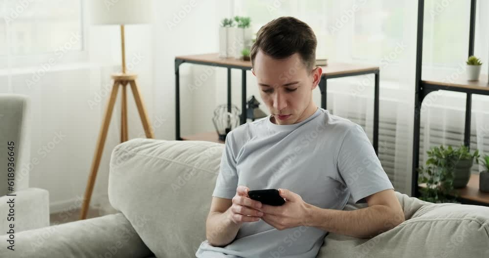 Contemplative Caucasian man sitting on a sofa in casual attire and engrossed in using his phone. He appears deep in thought, texting or browsing content, while enjoying the comfort of his home. Contemplative Caucasian man sitting on a sofa in casual attire and engrossed in using his phone. He appears deep in thought, texting or browsing content, while enjoying the comfort of his home.