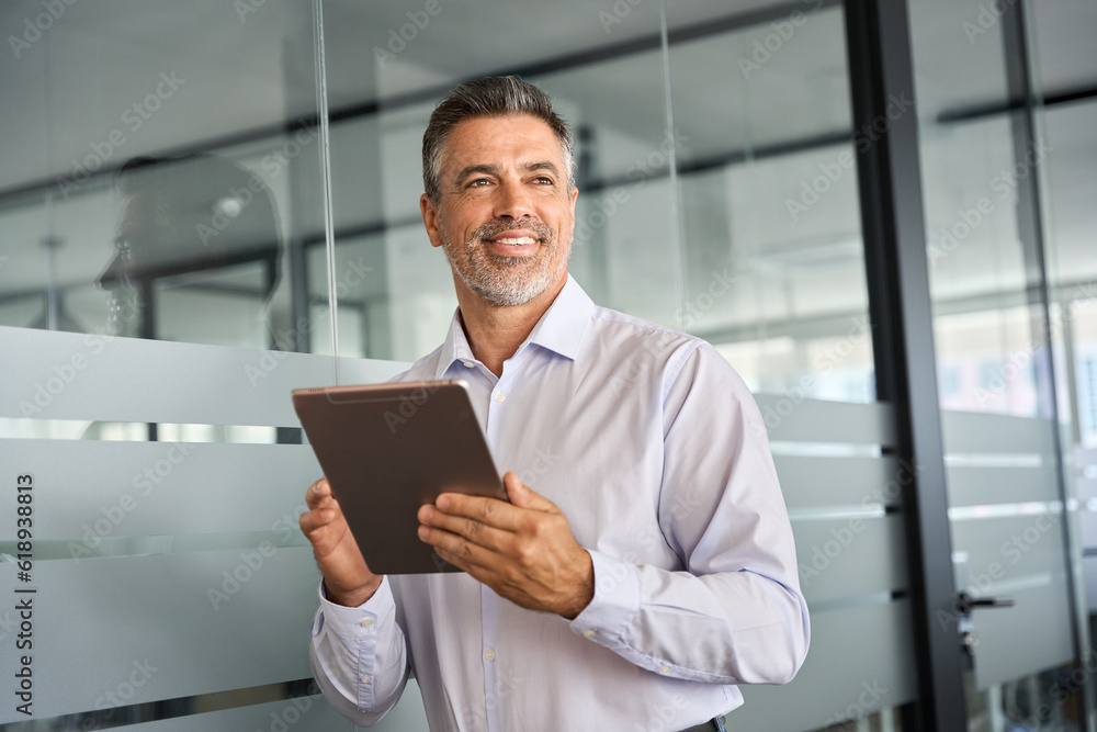 Fototapeta premium Happy middle aged business man ceo standing in office using digital tablet. Smiling mature businessman professional executive manager looking away thinking working on tab computer.