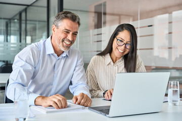 Happy smiling diverse colleagues executives team two professional managers looking at laptop pc having virtual meeting, watching webinar working together on online project sitting at office table.