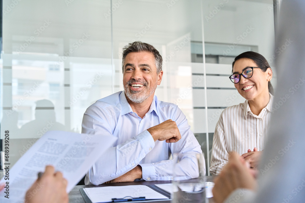 Happy diverse business people international corporate executive team talking at group meeting. Smiling older Latin male manager working with colleagues collaborating at boardroom table. Happy diverse business people international corporate executive team talking at group meeting. Smiling older Latin male manager working with colleagues collaborating at boardroom table.