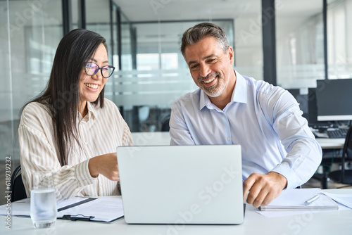 Two happy diverse professional colleagues Latin business man and Asian woman corporate executives working together in office using laptop, talking and looking at computer, having meeting discussion.