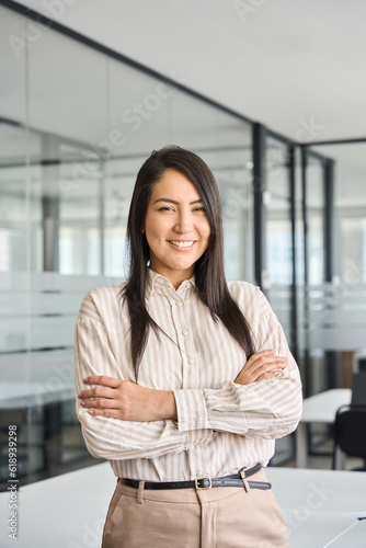 Confident smiling young professional Asian business woman corporate leader, happy japanese female employee, company sales marketing manager, standing arms crossed in office, vertical portrait.