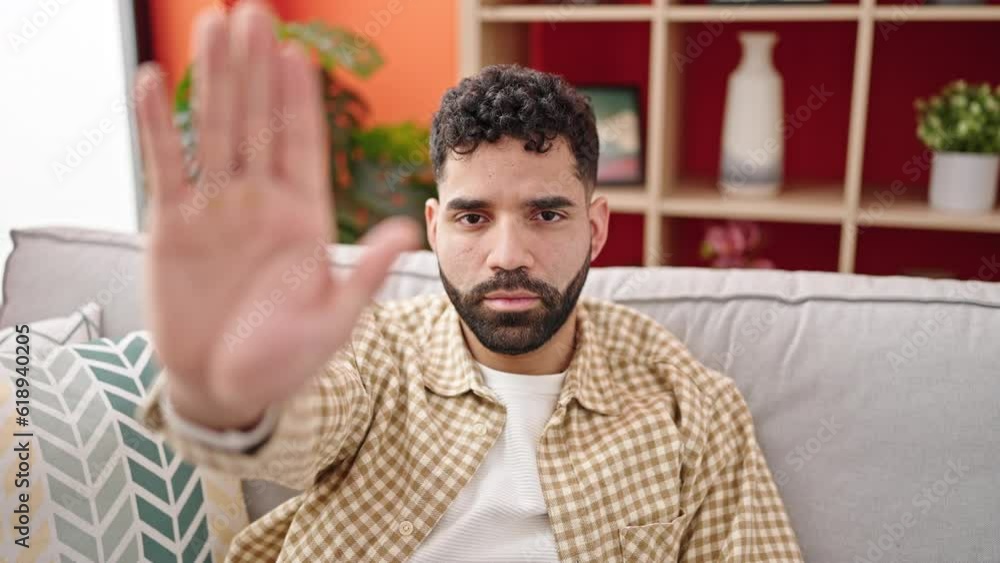 Young hispanic man sitting on sofa doing stop gesture at h Young hispanic man sitting on sofa doing stop gesture at h