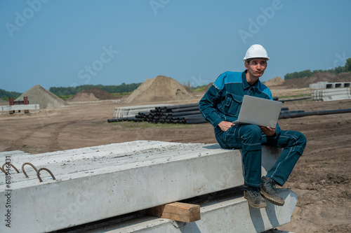Caucasian male builder in hardhat sits on floor slabs and uses laptop at construction site.  © Михаил Решетников